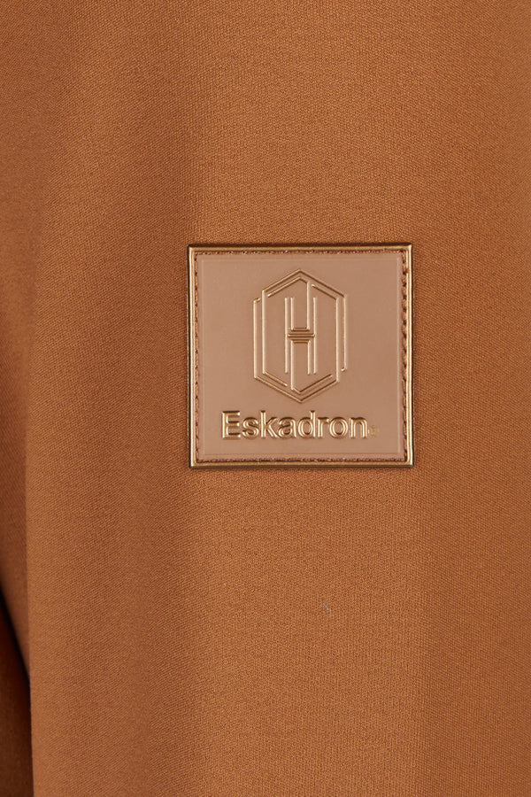 "Patch rectangulaire beige brodé sur un tissu marron, portant le logo géométrique en relief avec la lettre 'H' et le texte 'Eskadron'. "