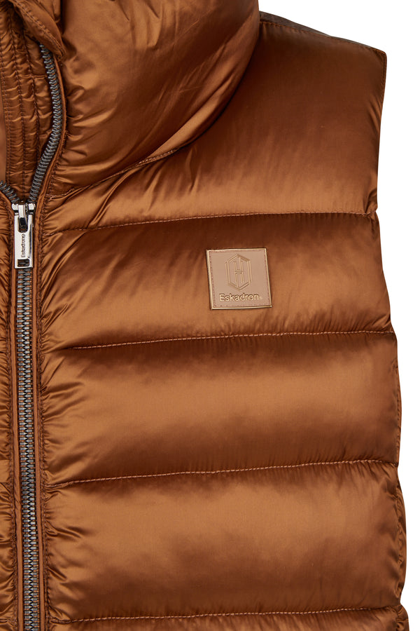 Gilet matelassé en tissu brillant marron caramel, avec une fermeture éclair argentée sur le côté gauche et le logo "Eskadron" en relief embossé sur un patch en cuir marron en haut à droite.