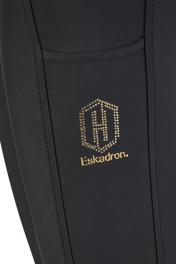 Balise alt : "Détail d'un vêtement noir avec un logo en strass dorés représentant un blason avec la lettre 'H'. Le nom de la marque 'Eskadron' est brodé en lettres dorées en dessous du logo."