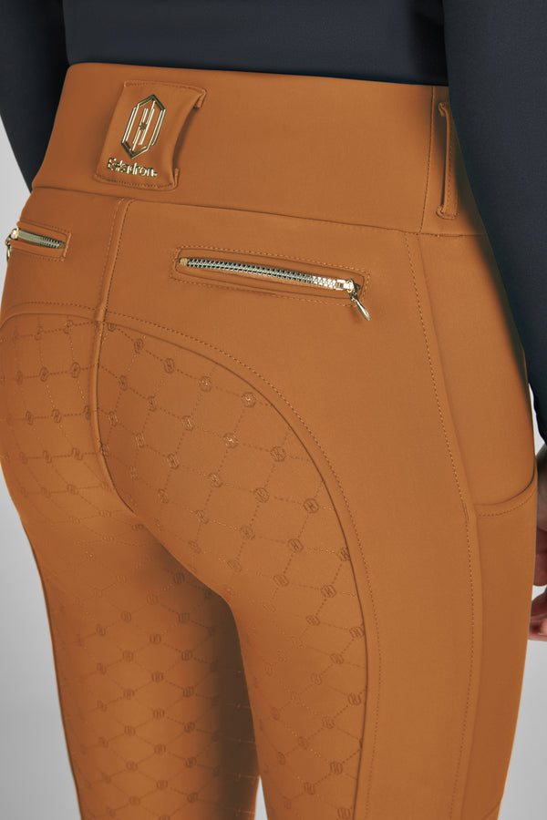 Vue de dos d'un pantalon d'équitation ajusté de couleur marron avec un motif géométrique à l'arrière et deux poches zippées avec fermeture dorée. Un logo métallique est visible sur la ceinture haute du pantalon. Le haut de la personne portant le pantalon est noir.