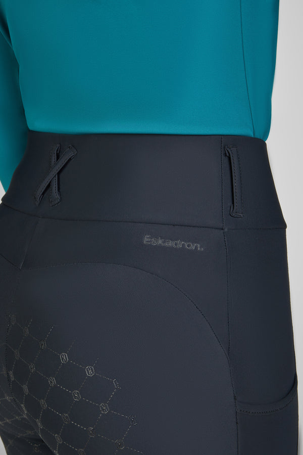 Vue rapprochée d'une personne portant un pantalon d'équitation bleu foncé avec la marque "Eskadron" embossée à l'arrière de la taille. Le pantalon présente des motifs géométriques sur la partie inférieure. La personne porte un haut turquoise.