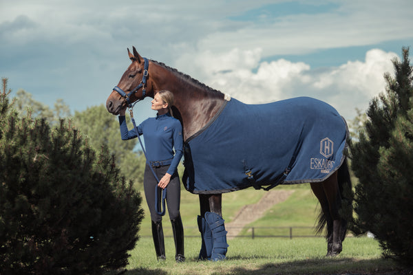 Une cavalière en tenue d'équitation bleu marine tient un cheval bai en licol, recouvert d'une couverture bleue avec le logo "Eskadron" sur le côté. Les deux sont debout dans un cadre naturel verdoyant sous un ciel nuageux. Le cheval porte également des protections sur ses pattes avant.