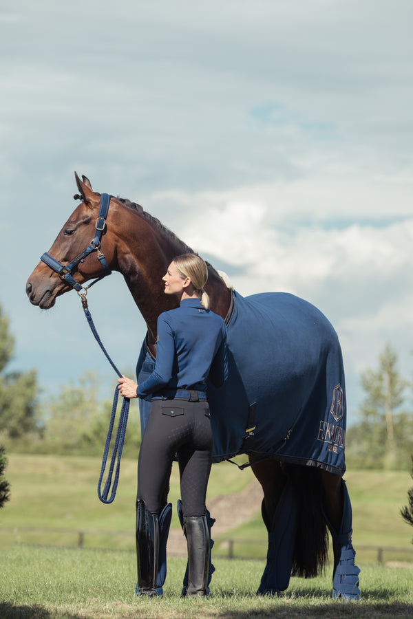 Une personne debout dans un champ, vêtue d'une tenue équestre bleue, tient un grand cheval brun portant une couverture et des protège-jambes assortis dans un paysage extérieur sous un ciel nuageux.