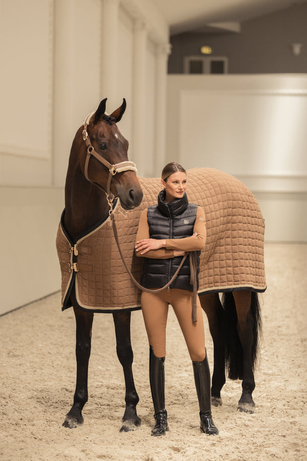 "Femme debout aux côtés d'un cheval brun portant une couverture de protection beige, dans une écurie lumineuse. La femme est vêtue d'une tenue d'équitation composée d'un gilet noir sans manches, d'un haut beige, de leggings assortis et de bottes d'équitation noires. Elle tient la bride du cheval tout en posant sereinement."