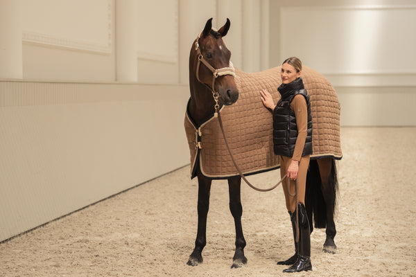 Une femme se tient à côté d'un cheval brun foncé couvert d'une couverture de protection marron, dans un manège intérieur. Elle porte une tenue équestre comprenant des bottes noires brillantes, un pantalon beige et une veste noire sans manche. La femme tient les rênes du cheval et pose une main sur lui dans un environnement calme et soigné avec un sol en sable.