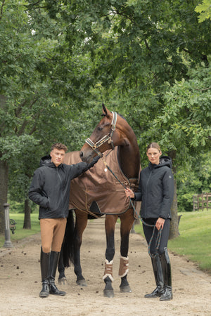 Deux personnes en vêtements d'équitation, un homme et une femme, tenant un cheval marron par une bride. Le cheval est vêtu d'une couverture de protection et porte des guêtres aux pattes avant. La scène se déroule sur une allée de sable, bordée d'arbres verts en arrière-plan.