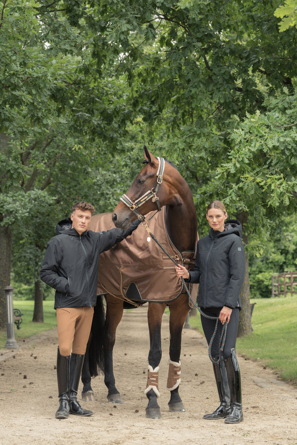 Deux personnes en vêtements d'équitation, un homme et une femme, tenant un cheval marron par une bride. Le cheval est vêtu d'une couverture de protection et porte des guêtres aux pattes avant. La scène se déroule sur une allée de sable, bordée d'arbres verts en arrière-plan.