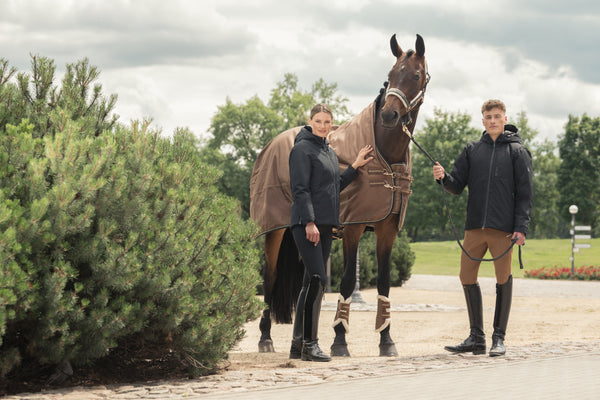 Alt : "Deux cavaliers habillés en noir posent avec un cheval portant une couverture marron dans un cadre extérieur verdoyant. Ils portent des bottes d'équitation noires, et le jeune homme tient les rênes du cheval."