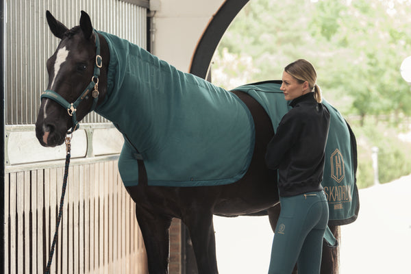 Femme prenant soin d'un cheval vêtu d'une couverture turquoise dans une écurie. La femme, habillée en tenue équestre assortie, est placée à côté du cheval, ajustant la couverture. L'écurie est moderne et lumineuse, avec une ouverture qui laisse entrevoir un environnement verdoyant à l'extérieur.