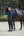 Une femme vêtue d'une veste matelassée bleue et de bottes d'équitation noires se tient à côté d'un cheval sombre équipé d'une selle et d'accessoires assortis en bleu. Le cheval porte également un bonnet bleu. La scène se déroule dans une arène d'équitation en plein air, avec des arbres verdoyants en arrière-plan.