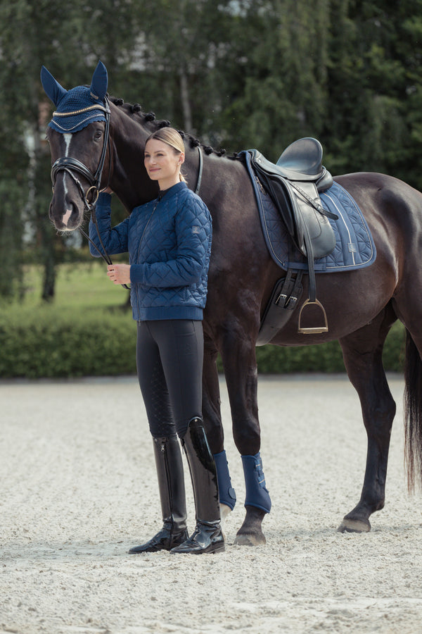 Une femme vêtue d'une veste matelassée bleue et de bottes d'équitation noires se tient à côté d'un cheval sombre équipé d'une selle et d'accessoires assortis en bleu. Le cheval porte également un bonnet bleu. La scène se déroule dans une arène d'équitation en plein air, avec des arbres verdoyants en arrière-plan.