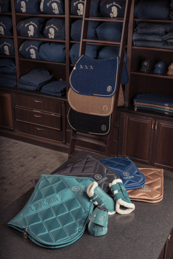 Image de plusieurs tapis de selle et protections pour chevaux de la marque Eskadron. Les tapis sont disposés sur une table et accrochés à une échelle, avec divers coloris tels que turquoise, bleu, beige et noir. En arrière-plan, des étagères présentent des équipements équestres soigneusement pliés, notamment des couvertures de selle avec des motifs de cheval brodé.