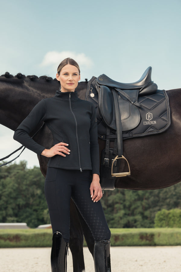 Femme habillée en tenue d'équitation noire, posant à côté d'un cheval noir portant une selle de marque Eskadron. Elle tient une main sur la hanche, avec une expression neutre. En arrière-plan, un paysage de verdure sous un ciel clair.