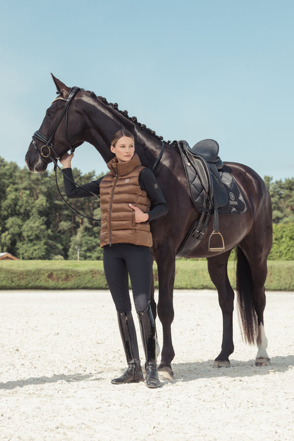 Une cavalière habillée d'une veste marron et d'un pantalon noir tient un cheval noir par la bride, se tenant à côté de lui. Le cheval porte une selle noire et a la crinière tressée. La scène se déroule en extérieur, sur un terrain sablonneux avec des arbres en arrière-plan, sous un ciel bleu clair.