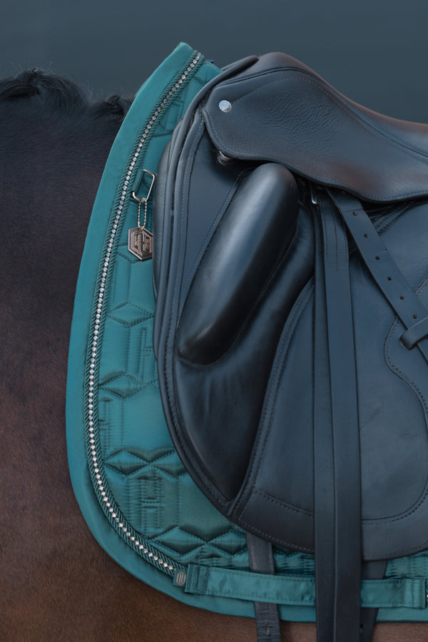 Gros plan d'une selle équestre noire posée sur un tapis de selle vert canard avec des motifs géométriques, attachée à un cheval brun. Un porte-clés métallique en forme d'hexagone pend à la bride du tapis de selle.