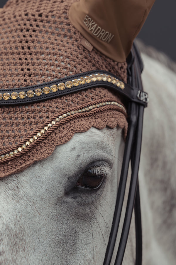 Gros plan sur la tête d'un cheval blanc portant un bonnet de protection d'oreilles de couleur marron, orné de strass dorés. Le bonnet est partiellement couvert par une bride en cuir noir décorée de pierres brillantes.