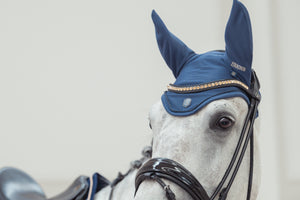 Cheval gris portant un bonnet anti-mouches bleu de la marque Eskadron, avec des détails dorés sur le front. Il porte également un mors et des rênes en cuir noir.