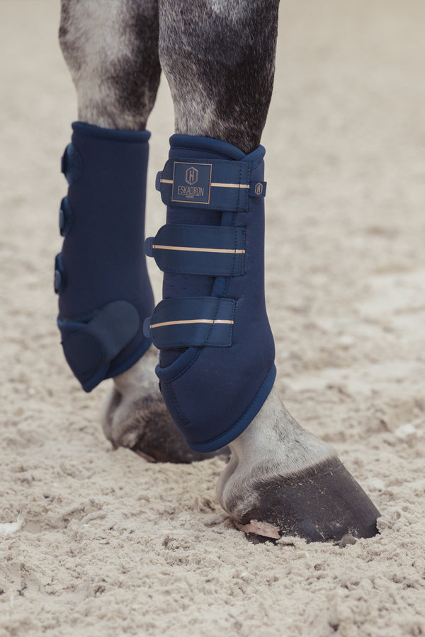 "Les jambes d'un cheval portant des guêtres de protection bleues de la marque Eskadron, attachées avec des sangles velcro. Le cheval se tient sur un sol sablonneux."