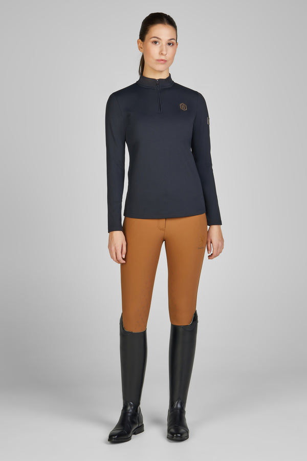 Femme portant une tenue d'équitation composée d'un haut à manches longues bleu sombre avec fermeture éclair partielle et d'un pantalon ajusté de couleur marron clair, accompagnée de bottes d'équitation noires montantes.