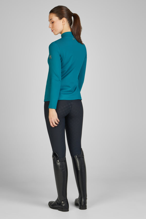 Femme de dos portant un haut turquoise à manches longues, un pantalon d'équitation noir et des bottes hautes en cuir noir, posant dans un studio au fond gris.