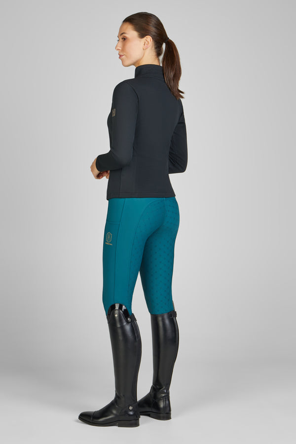 Femme vue de profil portant un pull noir moulant à col montant, des pantalons d'équitation bleus avec motifs texturés et des bottes hautes en cuir noir. Ses cheveux sont attachés en queue-de-cheval, et son expression est neutre.