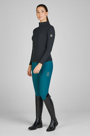 Femme posant, portant une tenue d'équitation composée d'une veste noire à fermeture éclair, un pantalon d'équitation turquoise avec des détails matelassés et le logo "Stübben" sur la cuisse, ainsi que des bottes d'équitation hautes en cuir noir. Fond gris uni.