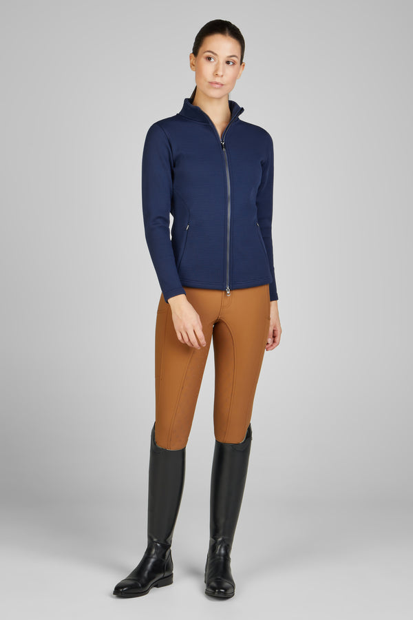 Femme portant une veste zippée bleu marine, un pantalon d'équitation marron et des bottes d'équitation noires, posant debout sur un fond gris neutre.