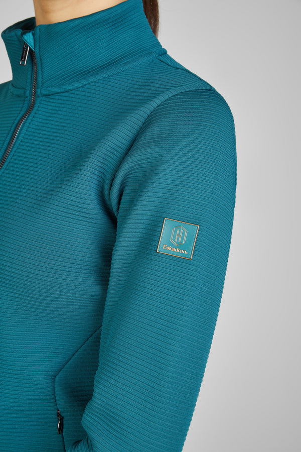 Alt : "Veste turquoise à texture côtelée avec une fermeture éclair partielle et un col montant. Un logo de la marque 'Eskadron' est visible sur la manche."