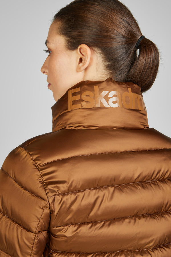 Femme de profil portant une veste matelassée marron avec le col relevé, où est inscrit "Eskadron" en lettres brillantes. Ses cheveux sont attachés en une queue de cheval basse.