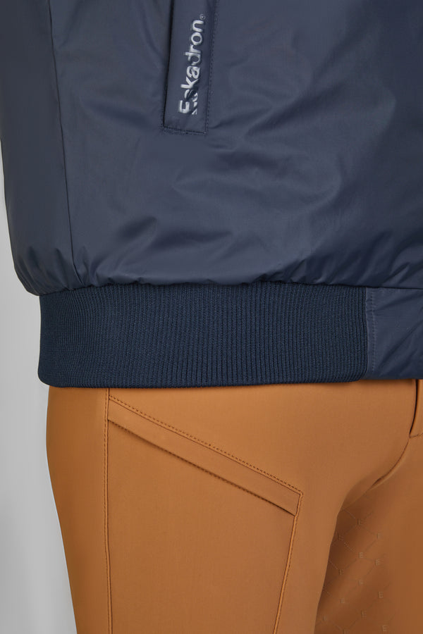 Gros plan sur une veste bleu marine avec un ourlet en bord-côte et un logo Eskadron visible à la verticale. La veste est associée à un pantalon ajusté de couleur marron camel.