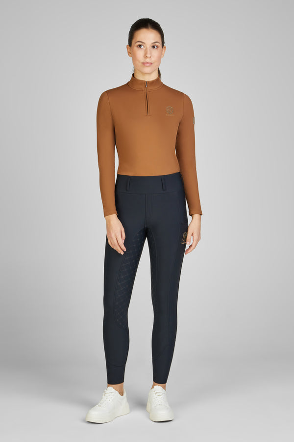 Femme debout portant un haut marron à manches longues avec un col zippé, un legging noir ajusté avec motifs discrets sur les jambes et des baskets blanches. Fond neutre.