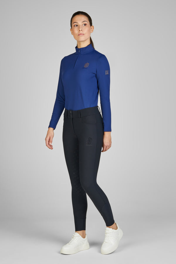 Femme portant une tenue de sport équestre composée d'un haut à manches longues bleu avec un col zippé, un legging noir ajusté avec des renforts sur les cuisses, et des baskets blanches.