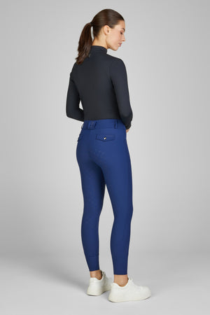 Femme de profil portant un haut à manches longues noir et un pantalon de sport bleu, avec des chaussures blanches. Elle a les cheveux attachés en queue de cheval et se tient debout sur un fond uni gris.