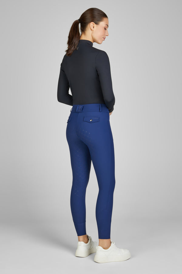 Femme de profil portant un haut à manches longues noir et un pantalon de sport bleu, avec des chaussures blanches. Elle a les cheveux attachés en queue de cheval et se tient debout sur un fond uni gris.