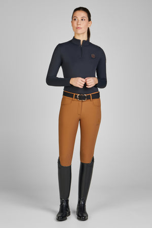 Femme debout portant un haut à manches longues bleu marine avec un col zippé, un pantalon d'équitation ajusté marron clair, une ceinture noire avec une boucle dorée, et des bottes d'équitation hautes et noires. Elle regarde vers le côté droit, les mains jointes devant elle sur un fond gris uni.