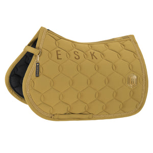 L'image présente un tapis de selle d'équitation de couleur jaune moutarde. Le tapis est conçu avec un motif matelassé géométrique composé de cercles et de lignes entrelacées, lui donnant une apparence texturée et élégante. Les bords du tapis sont finis avec un passepoil doré, ajoutant une touche de sophistication. Sur le côté droit du tapis, il y a un petit emblème doré en forme de hexagone avec un "H" stylisé au centre, probablement indiquant la marque du fabricant. Le tapis est également...
