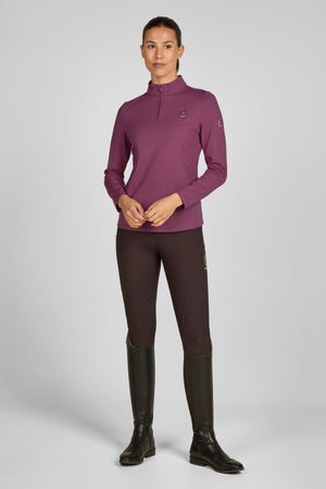 Dans cette image, une femme se tient debout, vêtue d'une tenue d'équitation. Elle porte un haut à manches longues de couleur bordeaux, qui possède une fermeture éclair partielle au niveau du col. Ce haut est ajusté et semble être en matière respirante, idéale pour les activités sportives. Elle porte également un pantalon d'équitation assorti, de couleur marron foncé, avec des surpiqûres et des détails contrastants. Le pantalon est conçu pour offrir confort et flexibilité tout en montant à...