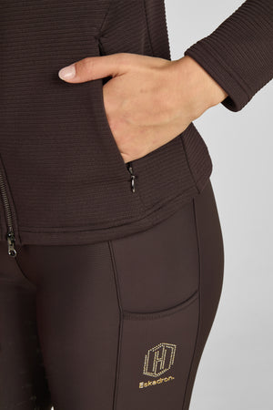 L'image montre une personne vêtue de vêtements de sport ou d'équitation, principalement de couleur marron. Voici une description détaillée : - **Haut du corps** : La personne porte un haut à manches longues avec une texture légèrement en relief, peut-être un tissu technique conçu pour l'exercice ou l'équitation. Le haut semble avoir un col montant, offrant une couverture complète du cou. - **Main** : La main de la personne est visible, avec les doigts légèrement pliés et posés sur la...