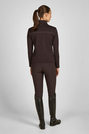 L'image présente une femme vue de dos, se tenant debout dans une posture droite. Elle porte une tenue d'équitation élégante et fonctionnelle. Voici une description détaillée : 1. **Haut** : La femme est vêtue d'une veste ou d'un haut à manches longues de couleur marron foncé. La veste semble être en tissu extensible, probablement conçue pour offrir confort et liberté de mouvement. Elle présente une fermeture éclair sur le devant et des détails en tissu contrastant sur les épaules et le haut...