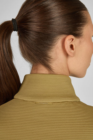 L'image montre une femme vue de côté et de dos. Ses cheveux sont attachés en une queue de cheval haute avec un élastique noir. Elle porte un vêtement à col haut, de couleur olive avec des rayures horizontales. Sur le col du vêtement, il y a un petit logo ou une étiquette cousue avec le texte "ISKO VITAL". La peau de la femme semble lisse et claire. L'image est prise sur un fond blanc uni, ce qui permet de bien voir les détails du vêtement et de la coiffure.