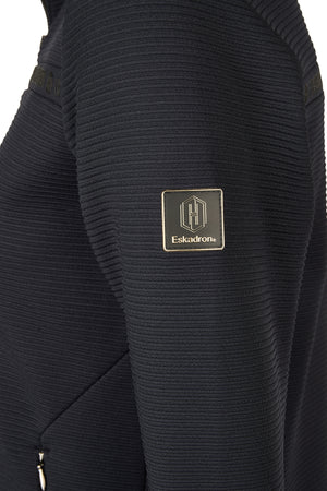 L'image montre une partie d'un vêtement, semblant être un pull ou un sweatshirt, de couleur noire. Le tissu présente un motif texturé avec des lignes horizontales régulières. Sur le côté gauche de l'image, on peut voir un patch rectangulaire avec des bordures dorées, portant le logo de la marque Eskadron. Le logo est composé d'une maison stylisée, accompagné du nom "Eskadron" inscrit en dessous en lettres capitales. Le patch se distingue par son aspect légèrement brillant et les bordures...