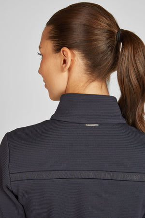 L'image montre une femme vue de dos, portant un vêtement gris foncé, probablement un haut à manches longues avec un col montant. Ses cheveux sont attachés en queue de cheval haute, maintenue par un élastique noir. Le tissu du haut semble texturé et strié, ce qui lui donne une apparence sportive ou athlétique. Sur le côté droit du haut, près du col, il y a une petite étiquette ou un logo avec le texte "Escapada" écrit dessus. De plus, il y a une bande horizontale en relief avec un motif...