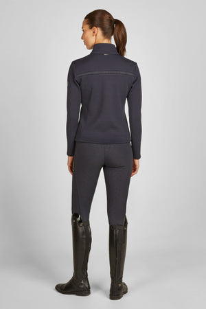 L'image montre une femme vue de dos, portant une tenue équitation moderne et élégante. Elle a les cheveux bruns rassemblés en queue de cheval haute. Voici une description détaillée de sa tenue : 1. **Haut** : Elle porte une veste ajustée bleu foncé avec une texture en relief. La veste semble être faite d'un matériau respirant, idéal pour les activités sportives. Elle a un col haut et des manches longues, et semble être légèrement rembourrée ou matelassée. La découpe est cintrée à la taille,...