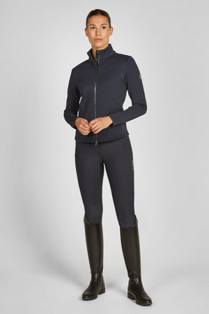Sur l'image, une personne est debout en position droite, les mains jointes devant elle. Elle porte une tenue d'équitation moderne et élégante. Elle est vêtue d'une veste ajustée de couleur bleu foncé, zippée jusqu'au cou, avec des détails texturés sur le devant et les manches. La veste semble offrir une bonne isolation thermique et un confort optimal pour les activités en extérieur. La partie inférieure de sa tenue comprend un pantalon d'équitation également bleu foncé, qui épouse...