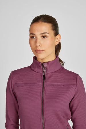 L'image montre une femme vêtue d'une veste de sport violette. Voici une description détaillée : 1. **La Femme** : - Elle a les cheveux tirés en arrière dans une queue de cheval serrée. - Elle a un teint clair, avec un maquillage léger et naturel. - Son expression faciale est neutre et elle regarde directement la caméra. 2. **La Veste** : - La veste est violette avec une texture fine et distincte. - Elle est fermée par une fermeture éclair centrale noire qui monte jusqu'au...