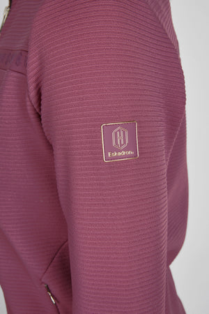 L'image montre un vêtement de sport en matière texturée de couleur rose. Le tissu semble être doux et extensible, probablement adapté pour des activités sportives ou de plein air. Sur la manche, on peut apercevoir un logo distinctif. Ce logo est de forme rectangulaire avec des angles arrondis. À l'intérieur du rectangle, il y a un emblème composé d'un "H" stylisé au centre, accompagné du texte "Eskadron" en dessous. L'ensemble du logo est réalisé en couleur or, offrant un contraste frappant...