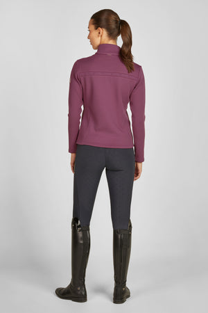 L'image montre une femme vue de dos. Elle porte une tenue qui semble adaptée pour l'équitation ou des activités sportives de plein air. Voici une description détaillée : 1. **Haut** : - **Couleur** : Le haut est de couleur violette. - **Coupe** : Il a une coupe ajustée avec des manches longues. - **Détails** : Il y a des coutures horizontales sur le tissu, donnant un effet texturé. La coupe est légèrement cintrée au niveau de la taille. - **Fermeture** : On peut voir une...