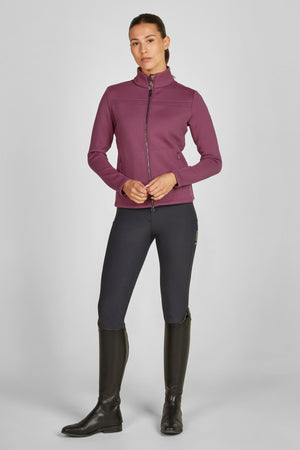 L'image montre une femme se tenant debout contre un fond blanc uni. Elle porte une tenue adaptée à l'équitation ou à une activité sportive similaire. Voici une description détaillée de sa tenue : 1. **Veste** : Elle porte une veste ajustée de couleur bordeaux. La veste est apparemment faite d'un tissu technique, probablement pour offrir confort et flexibilité. Elle a un col montant et une fermeture éclair complète à l'avant. La veste présente des coutures verticales et horizontales qui lui...