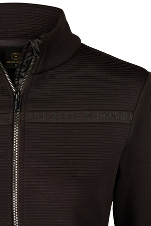 L'image montre une veste noire à fermeture éclair. La veste présente un design élégant et moderne avec une texture légèrement nervurée visible sur toute sa surface. Elle possède un col montant qui ajoute une touche de sophistication. La fermeture éclair centrale est de couleur foncée, probablement noire, et s'étend du bas de la veste jusqu'au col. De chaque côté de la fermeture éclair, il y a une bande horizontale noire avec le mot "Eskadron" répété en lettres blanches. Cette bande semble...