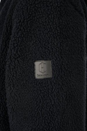 L'image montre un tissu noir et moelleux, probablement en laine ou en polaire. Au centre de l'image, il y a un badge carré avec des coins arrondis. Le badge est noir et argenté, et il est fixé au tissu. Le badge porte le logo de la marque "Eskadron" avec un symbole stylisé "E" en haut et le mot "Eskadron" écrit en dessous. Le tissu semble être une partie d'un vêtement, peut-être un manteau ou une veste, et le badge est probablement une étiquette de marque ou un logo.